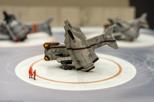 A futuristic spacecraft model is placed on a display stand with two miniature figures in orange suits nearby. The background features two additional spacecraft models, blurred to emphasize the one in the foreground.