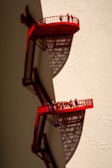 A surreal artwork featuring two small, red observation platforms with railings, attached to a beige wall. Tiny figures of people are standing on the platforms, casting intricate shadows below that add to the complexity of the scene.