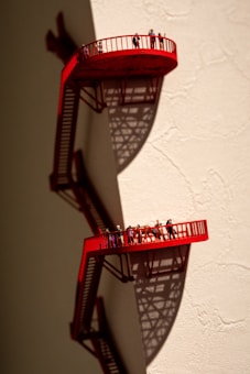 A surreal artwork featuring two small, red observation platforms with railings, attached to a beige wall. Tiny figures of people are standing on the platforms, casting intricate shadows below that add to the complexity of the scene.