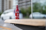 A red Coca-Cola bottle with branding and Japanese characters is placed on a wooden bench. The background is blurred, featuring a white car and green foliage in an urban setting.