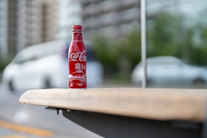 A red Coca-Cola bottle with branding and Japanese characters is placed on a wooden bench. The background is blurred, featuring a white car and green foliage in an urban setting.
