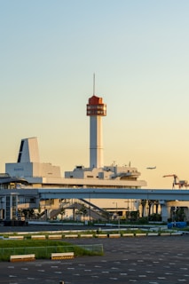 A large industrial or transportation complex is in the foreground, with a tall tower featuring an orange top. An airplane flies in the distance, and cranes are visible near the horizon, suggesting proximity to a port or shipping area. The area is lit by the warm glow of the setting or rising sun, casting soft yellow and golden hues.