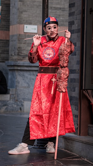 A person is wearing traditional Asian attire, including a bright red robe with intricate designs, a hat, and round sunglasses. They are holding a large skewer of food and have a playful expression.