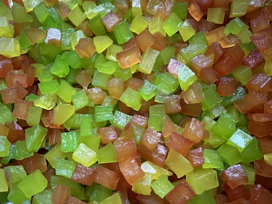 A close-up view of numerous small, translucent cubed pieces that appear to be candied fruit or jelly. The cubes are predominantly in shades of green and orange, with a shiny surface that reflects light.