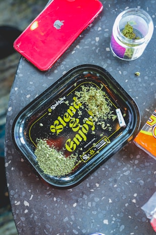 A red smartphone with an Apple logo rests on a speckled surface next to a plastic tray displaying the words 'Blend & Enjoy' in yellow lettering. The tray contains a small amount of green herbal material. Beside the tray, there is an open jar containing similar green material.