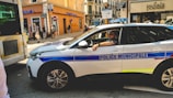 A municipal police car is parked on a busy city street, with a police officer sitting inside. The street is lined with various shops, including a Crédit Mutuel and another shop named Sauvage Poesie. Pedestrians are walking on the sidewalk, and there is a partially visible bus or vehicle on the left. The scene depicts a typical urban environment with a mix of vehicles and pedestrians.
