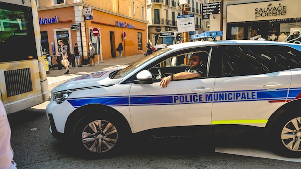 A municipal police car is parked on a busy city street, with a police officer sitting inside. The street is lined with various shops, including a Crédit Mutuel and another shop named Sauvage Poesie. Pedestrians are walking on the sidewalk, and there is a partially visible bus or vehicle on the left. The scene depicts a typical urban environment with a mix of vehicles and pedestrians.