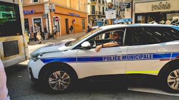 A municipal police car is parked on a busy city street, with a police officer sitting inside. The street is lined with various shops, including a Cr&eacute;dit Mutuel and another shop named Sauvage Poesie. Pedestrians are walking on the sidewalk, and there is a partially visible bus or vehicle on the left. The scene depicts a typical urban environment with a mix of vehicles and pedestrians.