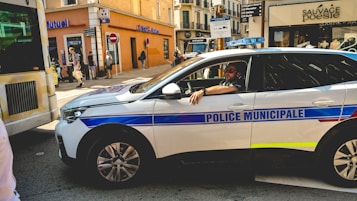 A municipal police car is parked on a busy city street, with a police officer sitting inside. The street is lined with various shops, including a Cr&eacute;dit Mutuel and another shop named Sauvage Poesie. Pedestrians are walking on the sidewalk, and there is a partially visible bus or vehicle on the left. The scene depicts a typical urban environment with a mix of vehicles and pedestrians.