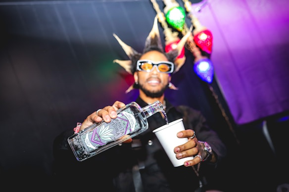 A person with spiked hair and sunglasses is pouring a clear liquid from a uniquely designed bottle into a white cup. Bright colored lights, including green, red, and blue, illuminate the background, enhancing the vibrant feel of the image.