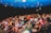 A group of people are seated in a theater, attentively facing the stage. Some seats are marked as 'VIP' in the middle row, indicating a special section. The audience appears diverse, with individuals wearing casual attire. The theater has a blue ceiling with lights, creating a vibrant atmosphere.
