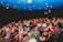 A group of people are seated in a theater, attentively facing the stage. Some seats are marked as 'VIP' in the middle row, indicating a special section. The audience appears diverse, with individuals wearing casual attire. The theater has a blue ceiling with lights, creating a vibrant atmosphere.