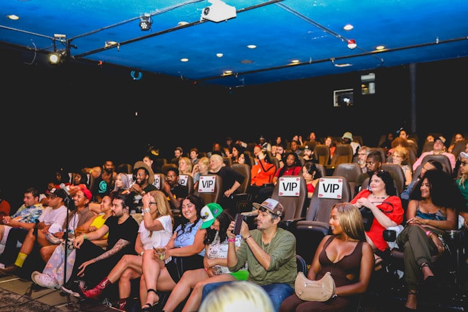 A group of people are seated in a theater, attentively facing the stage. Some seats are marked as 'VIP' in the middle row, indicating a special section. The audience appears diverse, with individuals wearing casual attire. The theater has a blue ceiling with lights, creating a vibrant atmosphere.