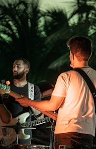 Two men are playing guitars in an outdoor setting. The scene is surrounded by what appears to be tropical foliage, and the lighting suggests it could be during sunset or under artificial lights. One man is wearing a white shirt while the other is in a black shirt.