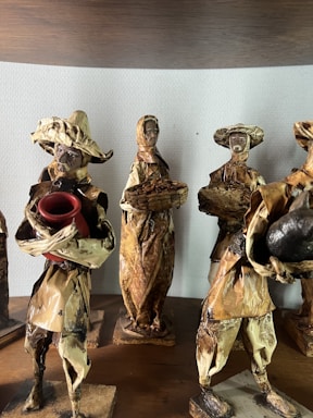 The image depicts a group of handcrafted figurines made from natural materials like dried leaves or bark. Each figure is dressed in rustic attire typical of traditional or folk settings. One holds a red pot, while another carries a basket. Their features are simple, adding to a rustic, artisanal charm.