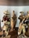 The image depicts a group of handcrafted figurines made from natural materials like dried leaves or bark. Each figure is dressed in rustic attire typical of traditional or folk settings. One holds a red pot, while another carries a basket. Their features are simple, adding to a rustic, artisanal charm.