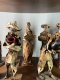 The image depicts a group of handcrafted figurines made from natural materials like dried leaves or bark. Each figure is dressed in rustic attire typical of traditional or folk settings. One holds a red pot, while another carries a basket. Their features are simple, adding to a rustic, artisanal charm.