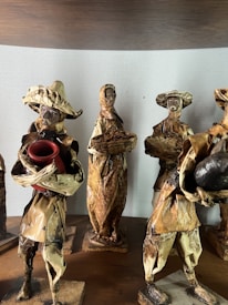 The image depicts a group of handcrafted figurines made from natural materials like dried leaves or bark. Each figure is dressed in rustic attire typical of traditional or folk settings. One holds a red pot, while another carries a basket. Their features are simple, adding to a rustic, artisanal charm.