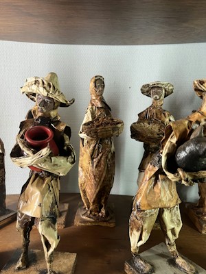 The image depicts a group of handcrafted figurines made from natural materials like dried leaves or bark. Each figure is dressed in rustic attire typical of traditional or folk settings. One holds a red pot, while another carries a basket. Their features are simple, adding to a rustic, artisanal charm.