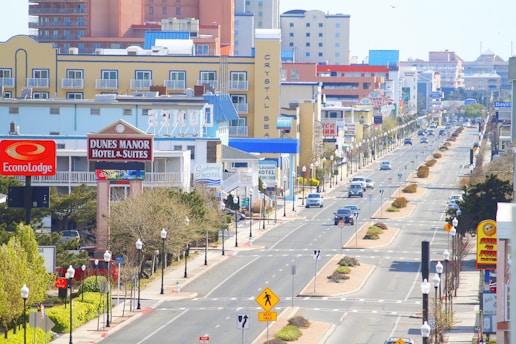 An urban street scene with various hotels and motels lining both sides. The street has several vehicle lanes and is adorned with street lamps and pedestrian crossing signs. Buildings display colorful signage, some of which are identifiable brands. Cars are driving and parked along the street. Trees and shrubs are planted along the sidewalks and in the central median.
