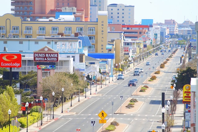 An urban street scene with various hotels and motels lining both sides. The street has several vehicle lanes and is adorned with street lamps and pedestrian crossing signs. Buildings display colorful signage, some of which are identifiable brands. Cars are driving and parked along the street. Trees and shrubs are planted along the sidewalks and in the central median.