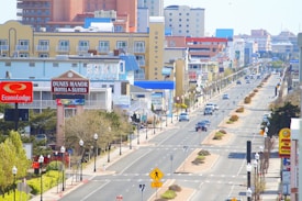 An urban street scene with various hotels and motels lining both sides. The street has several vehicle lanes and is adorned with street lamps and pedestrian crossing signs. Buildings display colorful signage, some of which are identifiable brands. Cars are driving and parked along the street. Trees and shrubs are planted along the sidewalks and in the central median.