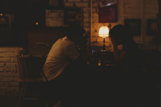 A dimly lit setting with two people sitting across from each other at a table. The warm glow of a small lamp on the table casts a soft light over their silhouettes. The background includes a brick wall with various framed items and decorations. The atmosphere suggests a quiet, intimate conversation.