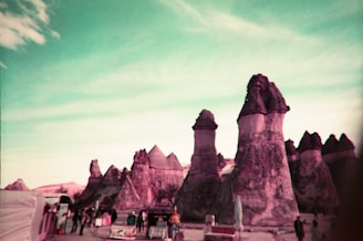 A surreal landscape features large, unusual rock formations resembling chimneys amidst a turquoise sky. Several people stand near what appears to be vending stalls or small shops, adding a human element to the scene.