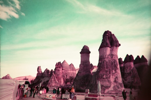 A surreal landscape features large, unusual rock formations resembling chimneys amidst a turquoise sky. Several people stand near what appears to be vending stalls or small shops, adding a human element to the scene.