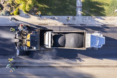 An aerial view of a road construction scene with workers in high-visibility vests guiding machinery as fresh asphalt is being laid down. A large truck is unloading asphalt onto the road surface, while a paving machine smooths and spreads the material. The setting is an urban street lined with grass and a sidewalk where multiple workers are coordinating the task.
