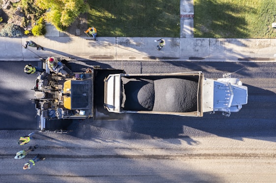 An aerial view of a road construction scene with workers in high-visibility vests guiding machinery as fresh asphalt is being laid down. A large truck is unloading asphalt onto the road surface, while a paving machine smooths and spreads the material. The setting is an urban street lined with grass and a sidewalk where multiple workers are coordinating the task.
