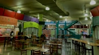 An indoor food court with multiple food stalls, including Taichi Asian Cuisine and Xich lo Viet Street Food. Several wooden tables and chairs are scattered around the area. A bright green column and teal staircase add color to the industrial ceiling with exposed pipes and hanging lights. Some people are seated at the tables, suggesting a relaxed dining atmosphere.