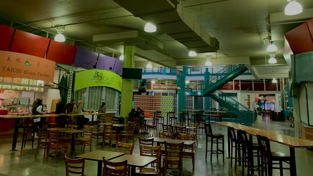 An indoor food court with multiple food stalls, including Taichi Asian Cuisine and Xich lo Viet Street Food. Several wooden tables and chairs are scattered around the area. A bright green column and teal staircase add color to the industrial ceiling with exposed pipes and hanging lights. Some people are seated at the tables, suggesting a relaxed dining atmosphere.