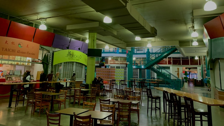 An indoor food court with multiple food stalls, including Taichi Asian Cuisine and Xich lo Viet Street Food. Several wooden tables and chairs are scattered around the area. A bright green column and teal staircase add color to the industrial ceiling with exposed pipes and hanging lights. Some people are seated at the tables, suggesting a relaxed dining atmosphere.