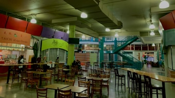 An indoor food court with multiple food stalls, including Taichi Asian Cuisine and Xich lo Viet Street Food. Several wooden tables and chairs are scattered around the area. A bright green column and teal staircase add color to the industrial ceiling with exposed pipes and hanging lights. Some people are seated at the tables, suggesting a relaxed dining atmosphere.