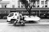 A flatbed truck filled with various items and produce is parked on the street, with a person sitting in the driver's seat looking out the window. In the foreground, a blurred motion of a rider on a motorcycle passes by swiftly. The background shows a building with multiple windows and a tree in front.