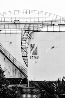 A large industrial storage tank with a distinctive spiral staircase and safety railings. Rust and weathering are visible on the surface, with a partially faded logo and text. Plants and a metal fence are in the foreground.