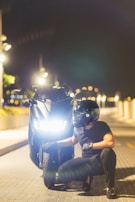 A person wearing a black helmet and gloves is crouching beside a black motorcycle on a dimly lit street at night. The motorcycle's headlight is on, illuminating the surroundings as streetlights glow softly in the background. The atmosphere is serene and slightly mysterious.