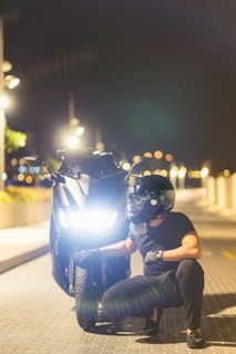 A person wearing a black helmet and gloves is crouching beside a black motorcycle on a dimly lit street at night. The motorcycle's headlight is on, illuminating the surroundings as streetlights glow softly in the background. The atmosphere is serene and slightly mysterious.