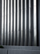 Tall vertical metal slats with a modern architectural design, featuring a series of evenly spaced horizontal lines, creating a sleek and industrial look. In the foreground, there are grey concrete panels at the base which contrast with the metallic texture above.