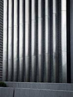 Tall vertical metal slats with a modern architectural design, featuring a series of evenly spaced horizontal lines, creating a sleek and industrial look. In the foreground, there are grey concrete panels at the base which contrast with the metallic texture above.