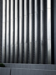 Tall vertical metal slats with a modern architectural design, featuring a series of evenly spaced horizontal lines, creating a sleek and industrial look. In the foreground, there are grey concrete panels at the base which contrast with the metallic texture above.