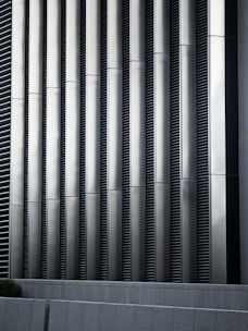 Tall vertical metal slats with a modern architectural design, featuring a series of evenly spaced horizontal lines, creating a sleek and industrial look. In the foreground, there are grey concrete panels at the base which contrast with the metallic texture above.