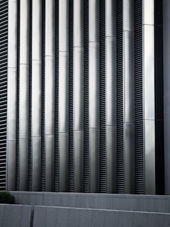 Tall vertical metal slats with a modern architectural design, featuring a series of evenly spaced horizontal lines, creating a sleek and industrial look. In the foreground, there are grey concrete panels at the base which contrast with the metallic texture above.
