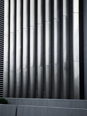 Tall vertical metal slats with a modern architectural design, featuring a series of evenly spaced horizontal lines, creating a sleek and industrial look. In the foreground, there are grey concrete panels at the base which contrast with the metallic texture above.