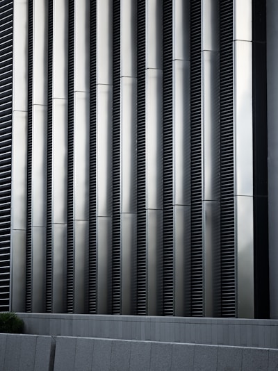 Tall vertical metal slats with a modern architectural design, featuring a series of evenly spaced horizontal lines, creating a sleek and industrial look. In the foreground, there are grey concrete panels at the base which contrast with the metallic texture above.