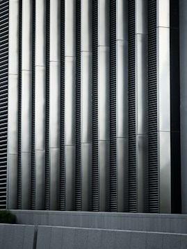 Tall vertical metal slats with a modern architectural design, featuring a series of evenly spaced horizontal lines, creating a sleek and industrial look. In the foreground, there are grey concrete panels at the base which contrast with the metallic texture above.