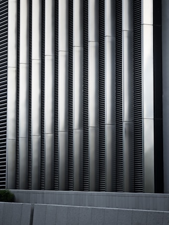Tall vertical metal slats with a modern architectural design, featuring a series of evenly spaced horizontal lines, creating a sleek and industrial look. In the foreground, there are grey concrete panels at the base which contrast with the metallic texture above.