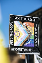 A protest sign is held up against a clear blue sky. The sign reads 'Fridays For Future' with additional phrases including 'Tax the Rich!' and 'Feed the Poor! Save the Planet!' There are colorful geometric shapes and a map illustration with the word 'Erlangen.' The sign suggests themes of climate action and social justice.