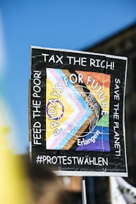 A protest sign is held up against a clear blue sky. The sign reads 'Fridays For Future' with additional phrases including 'Tax the Rich!' and 'Feed the Poor! Save the Planet!' There are colorful geometric shapes and a map illustration with the word 'Erlangen.' The sign suggests themes of climate action and social justice.